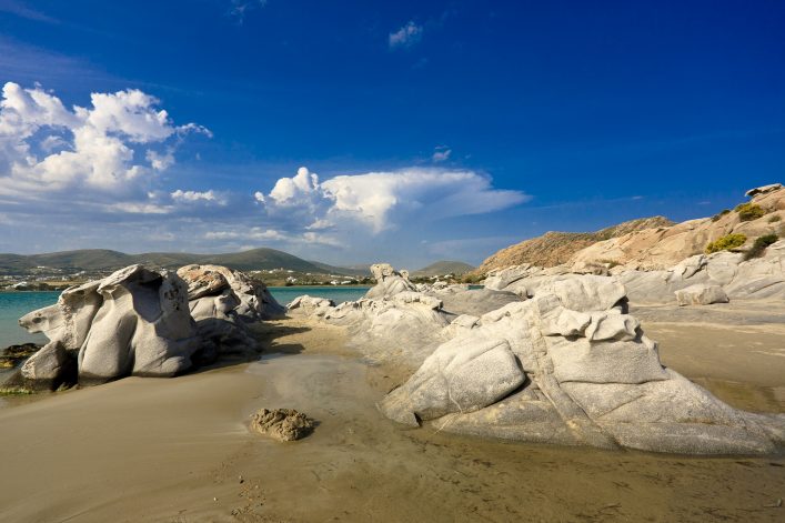 Weißgraue, glatt geschliffene Felsen ragen aus sandigem Boden bei Kolymbithres Beach, Paros. Im Hintergrund türkisfarbenes Meer, grüne Hügel und ein strahlend blauer Himmel mit weißen Wolken.