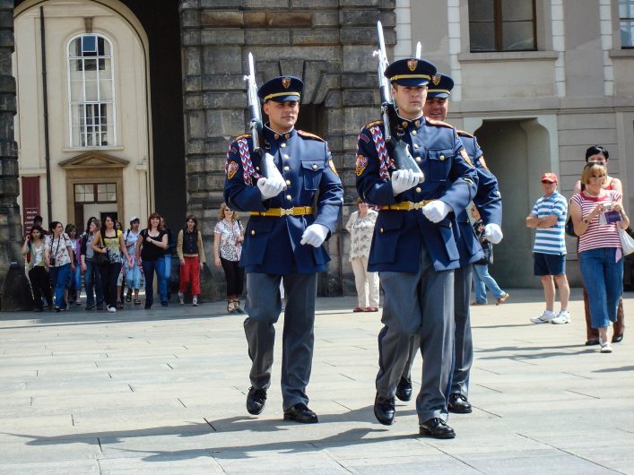 Drei Soldaten in blauen Uniformen marschieren synchron auf einem Platz, Gewehre geschultert. Im Hintergrund Gebäude mit Menschen, die zuschauen. Starker Sonnenschein beleuchtet die Szene.