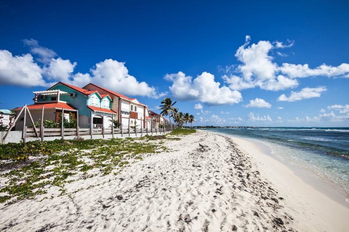 Strand mit feinem Sand, rechts das Meer. Links stehen bunte Häuser hinter einem Zaun, von Palmen gesäumt. Blauer Himmel mit wenigen Wolken. Vegetation am Strand verteilt.