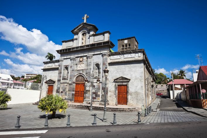 Kirchenfassade aus grauem Stein mit rotem Ziegeldach, zwei hölzerne Eingangstüren, darüber Statuen in Nischen. Ein Kreuz auf dem Dach. Rechteckiger Platz davor, Palmen links, blauer Himmel.