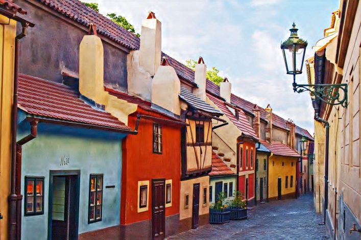 Bunte Häuser mit roten Dächern stehen dicht aneinander in der Goldenen Gasse, Prag. Links blaue und rote Fassaden, rechts eine Laterne an der Wand. Kopfsteinpflaster führt durch die enge, malerische Straße.