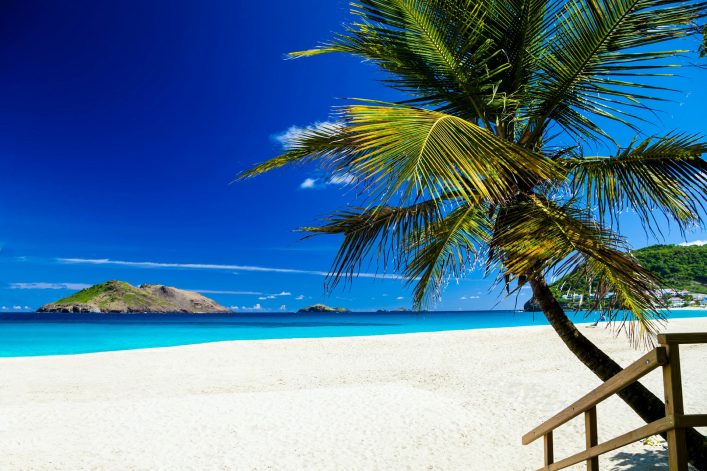 Strahlend blauer Himmel über einem tropischen Strand mit hellen Sand und türkisfarbenem Meer. Rechts sind Palmen und ein Holzgeländer sichtbar. Im Hintergrund eine grüne Insel im Meer.