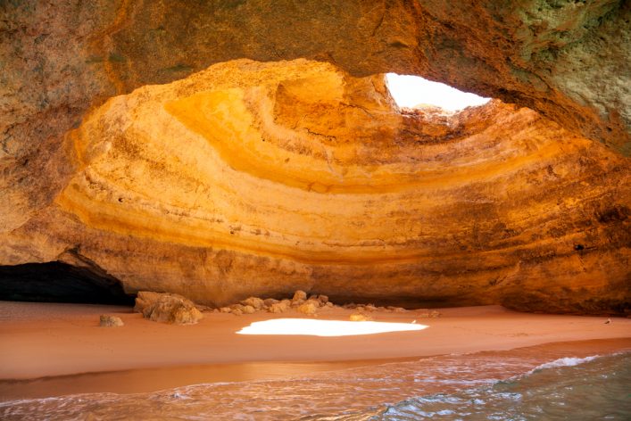 Sandstrand in einer Höhle mit natürlichem Licht von oben, das die Wände in warmen Gelb- und Orangetönen erleuchtet. Im Vordergrund sanftes Meereswasser, im Hintergrund Felsen und Schatten.