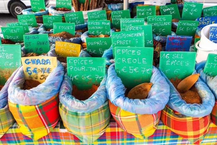 Gewürze in bunten Säcken auf einem Marktstand mit handgeschriebenen Schildern in Grün und Gelb, die Preise und Namen anzeigen. Die Säcke liegen dicht nebeneinander auf einem bunt karierten Tuch.