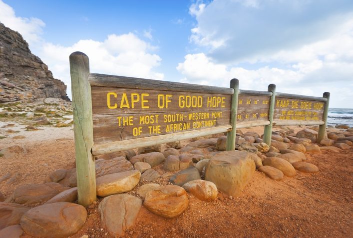 Holzschild am Kap der Guten Hoffnung, Südafrika, mit gelber Aufschrift über den südwestlichsten Punkt Afrikas, umgeben von Felsen und sandigem Boden, Meer im Hintergrund unter blauem Himmel.