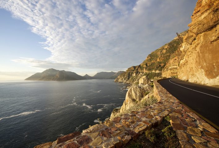 Küstenstraße am rechten Bildrand windet sich entlang einer steilen, felsigen Klippe. Links breitet sich das ruhige Meer aus, mit Bergen am Horizont unter einem teils wolkigen Himmel.