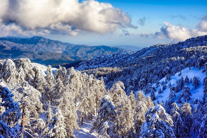 Verschneite Berge der Troodos-Gebirgskette mit zahlreichen Tannen, die mit Schnee bedeckt sind. Im Hintergrund majestätische Bergketten unter einem strahlend blauen Himmel mit dichten Wolken.