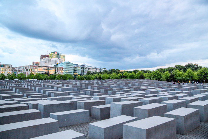 Graue Betonstelen in verschiedenen Höhen formen das Holocaust-Mahnmal in Berlin. Im Hintergrund lockere Baumreihe, darüber ein bedeckter Himmel. Rechts seitlich moderne Gebäude sichtbar.