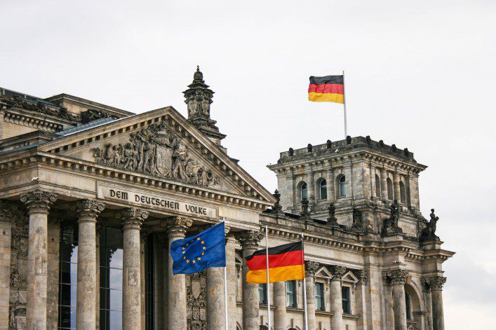 Reichstagsgebäude in Berlin mit historischer Fassade, Säulen und der Inschrift „Dem Deutschen Volke“. Vorne wehen die Europäische und zwei deutsche Flaggen. Oben eine weitere deutsche Flagge.