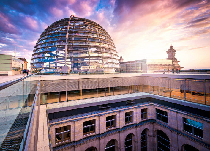 Gläserne Kuppel des Berliner Reichstags, umgeben von moderner Architektur, während der Himmel in rosa und lila Tönen schimmert. Eine Aussichtsplattform ist von Menschen besucht.