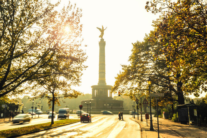 Siegessäule mit goldener Viktoria-Statue im Zentrum, von Sonnenlicht umgeben. Bäume rahmen die Szene links und rechts ein. Autos und Passanten auf der Straße im Vordergrund.