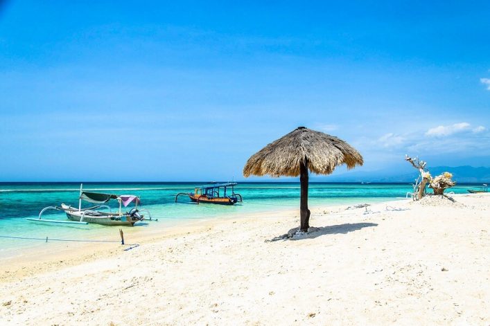 Strahlend weißer Sandstrand mit Strohdach-Sonnenschirm im Vordergrund, zwei Boote auf türkisblauem Wasser links und ein knorriger Baum im Hintergrund unter klarem, blauem Himmel.