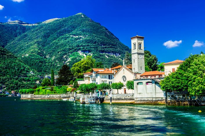 Kirche mit Uhrturm und roten Ziegeldächern, umgeben von grünen Bäumen, am Ufer des Lago di Como, links Berge im Hintergrund, blauem Himmel und wenigen Wolken darüber; ruhiges Wasser im Vordergrund.