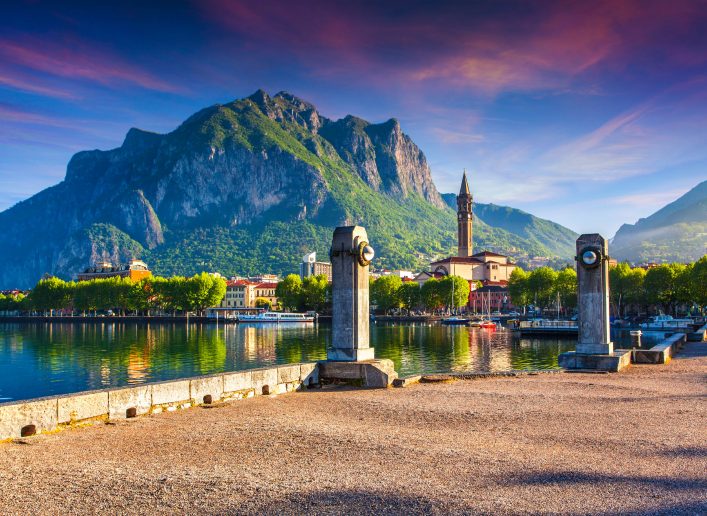 Malerisches Dorf am See mit bunten Häusern und Kirchturm im Vordergrund, eingebettet in grüne Berge. Drei Steinsäulen auf Uferpromenade, Abendhimmel in warmen Farben.