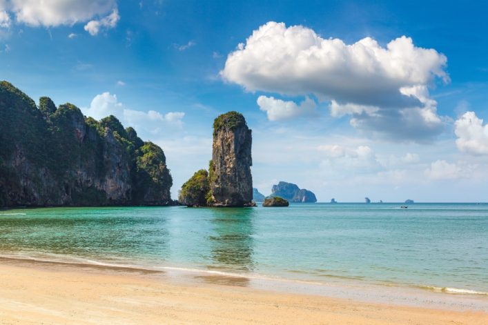 Weißer Sandstrand im Vordergrund, türkisblaues Meer und hohe, bewachsene Kalksteinfelsen im Hintergrund. Vereinzelte Wolken am blauen Himmel. Links die Küste, zentraler Felsen im Wasser.
