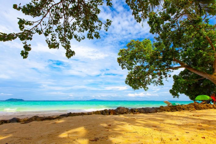 Strand mit goldenem Sand und grünen Bäumen am rechten Bildrand; türkisfarbenes Meer im Hintergrund, blauer Himmel mit Wolken; Steine und Liegestühle unter einem Sonnenschirm rechts.