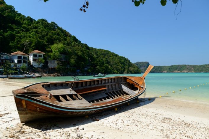 Ein traditionelles Holzboot liegt schräg am sandigen Strand. Links sind tropische Bäume und Gebäude, während rechts das türkisfarbene Wasser mit Bojen sichtbar ist. Der Himmel ist klar und blau.