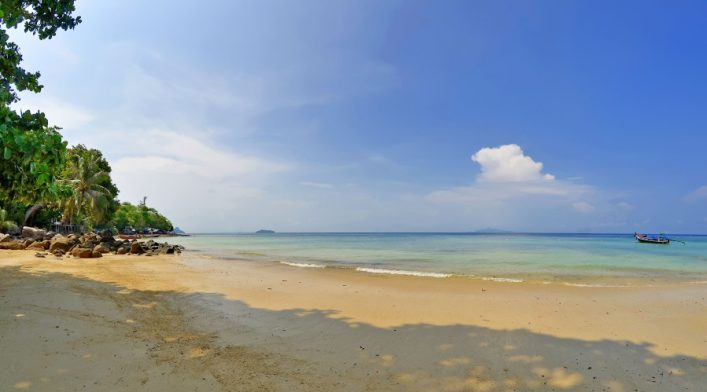 Sandstrand mit klarem Wasser, linken Rand von tropischen Bäumen und Felsen gesäumt. Ein Boot ankert rechts. Am Horizont eine Insel unter blauem Himmel mit weißen Wolken.