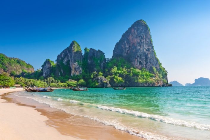 Goldener Sandstrand und türkisfarbenes Meer vor majestätischen Kalksteinfelsen, typisch für Thailands Küste, mit traditionellen Langbooten am Ufer.
