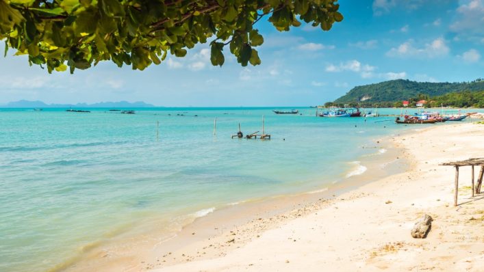 Am Thong Krut Strand leuchtet türkisblaues Wasser unter klarem Himmel. Boote schwimmen nahe der Küste, die von grünen Bäumen umrahmt ist. Der Sand verläuft sanft ins Wasser.