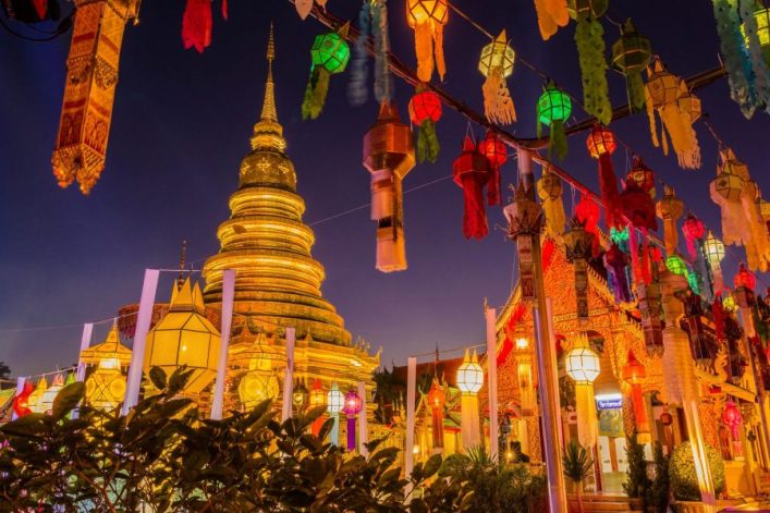 Goldene Pagode im Hintergrund, umgeben von bunten Laternen und Dekorationen in leuchtenden Farben bei Nacht. Strahlender Himmel, festliche Stimmung. Pflanzen im Vordergrund, Pagodendach leuchtet.
