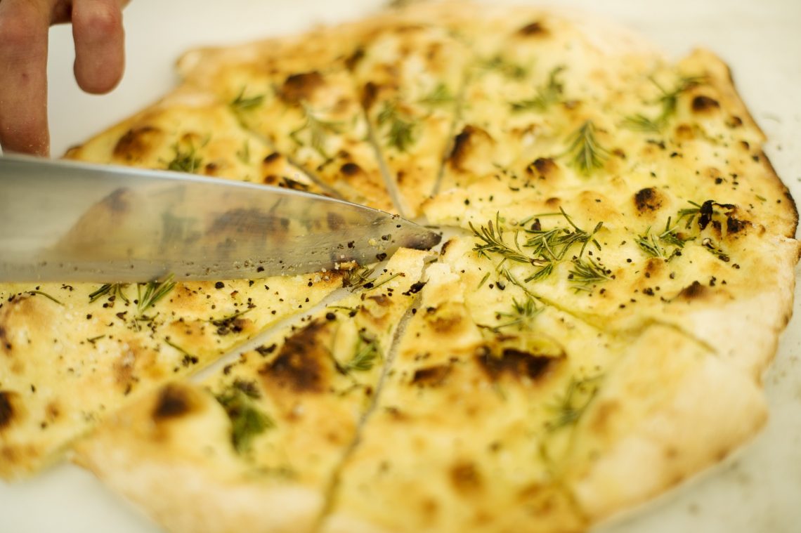 Flammkuchen mit goldbrauner Kruste wird mit einem Messer in Stücke geschnitten. Frische grüne Kräuter sind darüber gestreut. Das Messer befindet sich links im Bild.