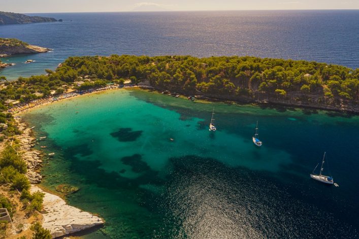 Strand von Aliki auf Thassos mit klarem türkisfarbenem Wasser. Links dichter Wald, rechts drei Segelboote auf dunklerem Wasser, im Hintergrund das weite Meer, Sonnenlicht schimmert auf der Oberfläche.