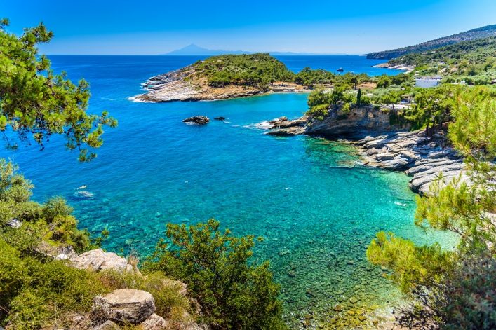 Eine türkisblaue Bucht mit klarem Wasser, umgeben von grünen Pinien und felsigen Klippen. Im Hintergrund erstreckt sich ein bewaldeter Hügel. Der Himmel ist wolkenlos und strahlend blau.