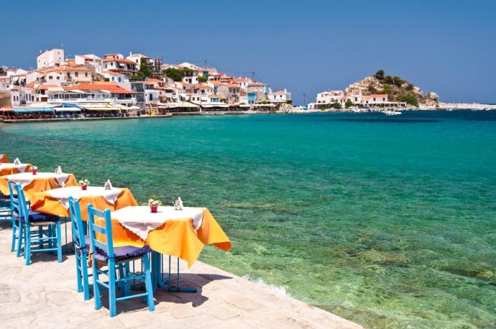 Türkisfarbenes Meer trifft auf eine Küste mit weißen Häusern und roten Dächern. Im Vordergrund stehen Tische mit orangefarbenen Tischtüchern und blauen Stühlen entlang der Promenade.