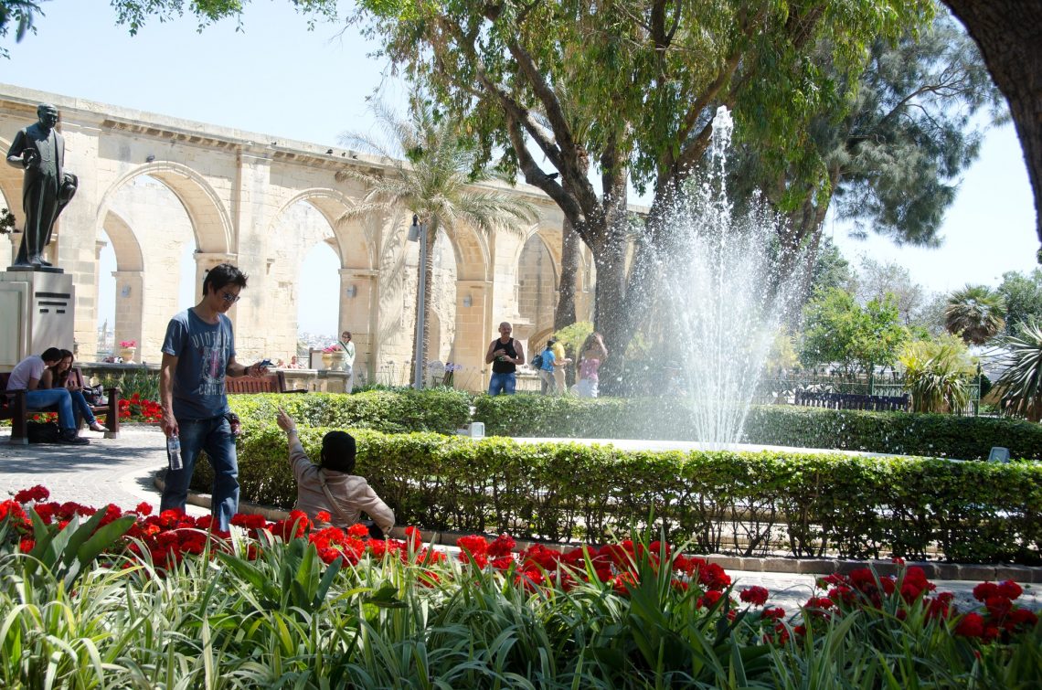 Im Upper Barrakka Garden sprudelt ein Brunnen in der Mitte, umgeben von roten Blumen und Büschen. Menschen spazieren, Bäume spenden Schatten, und im Hintergrund stehen alte Steinbögen.