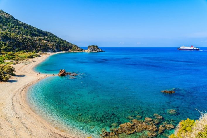 Azurblaues Meer an Potami Beach auf Samos, Griechenland. Links Kiesstrand mit grünen Bäumen, rechts oben fährt ein großes Schiff am Horizont. Klare, sonnige Atmosphäre.