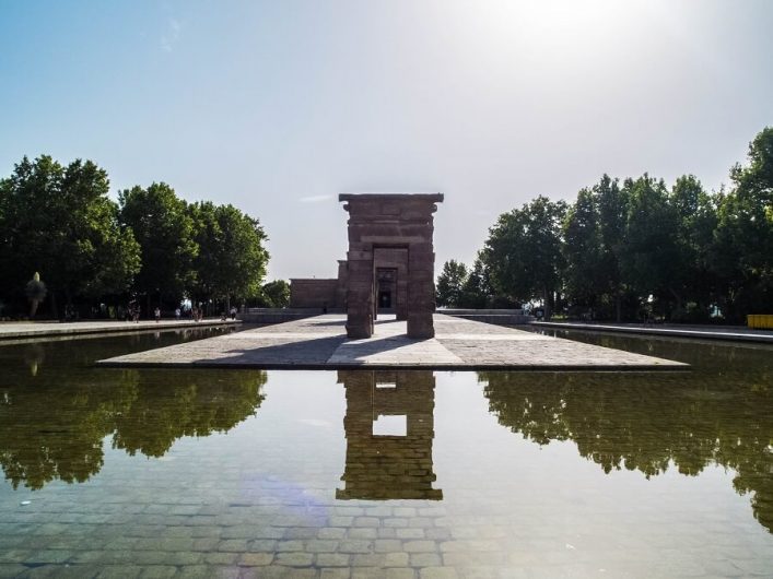 Ein antiker Tempel aus Stein thront zentral auf einer Plattform, umgeben von einem Wasserbecken. Links und rechts säumen Bäume den Hintergrund, während strahlendes Sonnenlicht von oben einfällt.