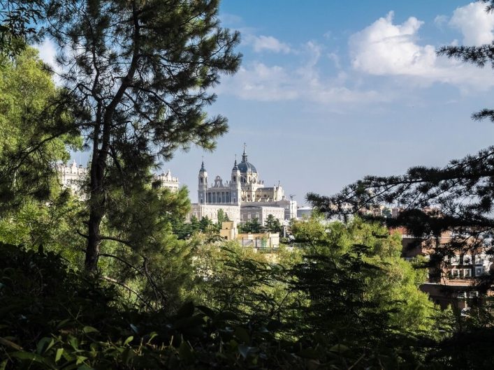 Im Vordergrund hohe Bäume und Büsche, die einen Blick auf eine majestätische Kathedrale mit zwei Türmen und einer Kuppel im Hintergrund freigeben. Der Himmel ist blau mit leichten Wolken.