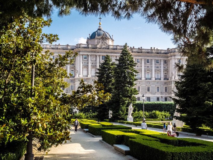 Majestätisches Gebäude mit Kuppel im Hintergrund, flankiert von hohen Bäumen. Im Vordergrund ein formaler Garten mit Hecken und Statuen. Besucher spazieren auf sandigen Wegen unter einem sonnigen Himmel.