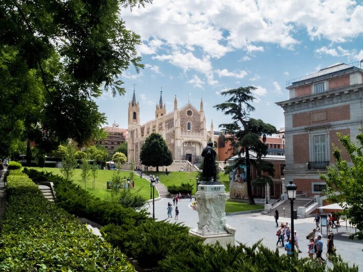 Die gotische Kirche mit spitzen Türmen steht im Hintergrund einer grünen Parklandschaft, eine Statue im Vordergrund. Rechts ist ein historisches Gebäude, während Menschen auf Wegen flanieren.