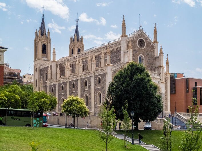 Gotische Kirche mit spitzen Türmen und Fenstern, umgeben von grünen Bäumen und Rasen. Im Vordergrund ein grasbewachsener Hang, Menschen spazieren, im Hintergrund ein blauer Himmel.