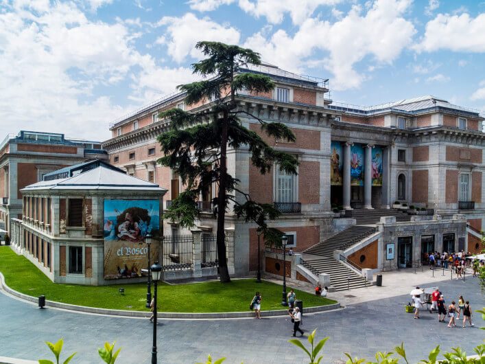 Großer, rechteckiger Backsteinbau mit Säulen und Treppen in der Mitte. Links eine Anzeigetafel mit Kunstmotiv. Vor dem Gebäude ein großer Baum und Menschen auf gepflastertem Platz bei bewölktem Himmel.
