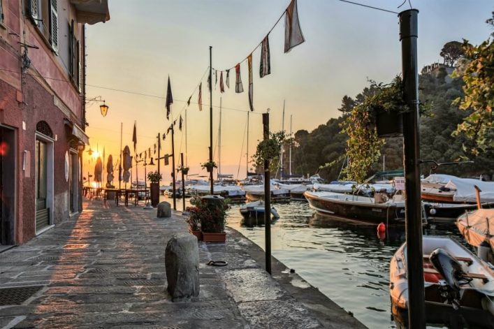 Sonnenuntergang über einem Hafen mit Booten rechts, ein gepflasterter Weg links neben einem Gebäude. Girlanden und Laternen hängen über dem Weg, Pflanzen schmücken ihn. Bäume und Hügel im Hintergrund.