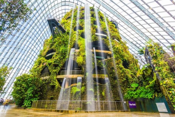 Hohe, begrünte Struktur im Glashaus mit großem Wasserfall; Pflanzen und Gehwege sind um die zentrale Säule verteilt. Stahlträger und Glasdach bilden die Kuppel, helle Atmosphäre inmitten der Natur.