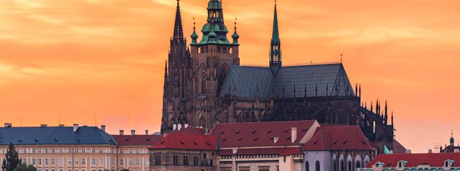 Majestätische Burg mit gotischen Türmen im Zentrum, umgeben von historischen Gebäuden mit roten Dächern. Der Himmel leuchtet orange beim Sonnenuntergang, und leichtes Grün ist unten sichtbar.
