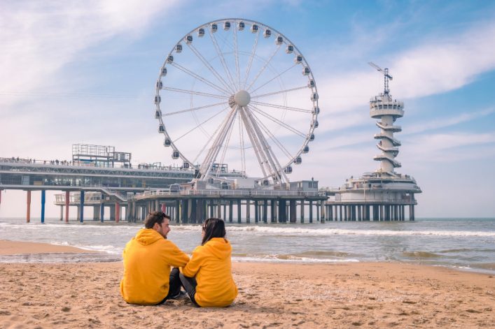 Schöner Strand bei Scheveningen: Im Vordergrund ein Paar in gelben Jacken auf Sand, im Hintergrund ein großes Riesenrad auf einem Pier, daneben ein markanter Turm, Himmel mit leichten Wolken.