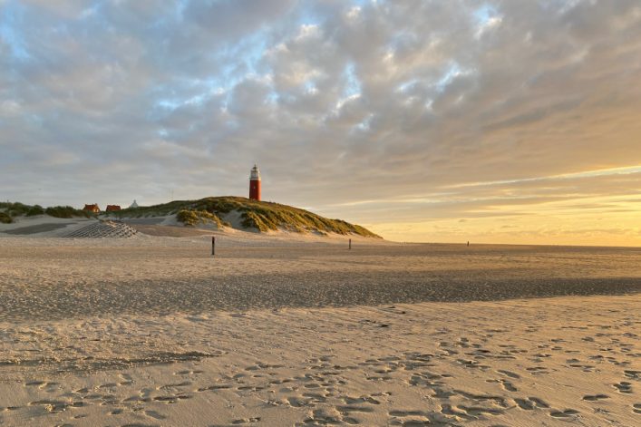 Sandstrand mit Fußspuren im Vordergrund, dahinter grüne Dünen. Ein roter Leuchtturm steht auf der Düne, von der Abendsonne beleuchtet. Im Hintergrund ist der Himmel bewölkt, löst sich jedoch in Goldtönen auf.