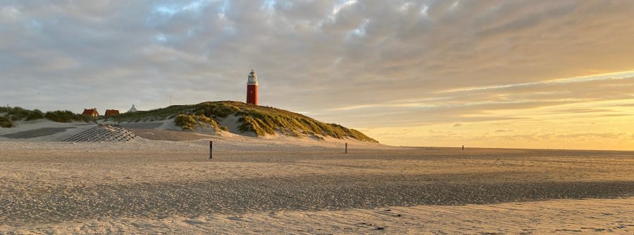 Sandstrand mit Fußspuren im Vordergrund, dahinter grüne Dünen. Ein roter Leuchtturm steht auf der Düne, von der Abendsonne beleuchtet. Im Hintergrund ist der Himmel bewölkt, löst sich jedoch in Goldtönen auf.