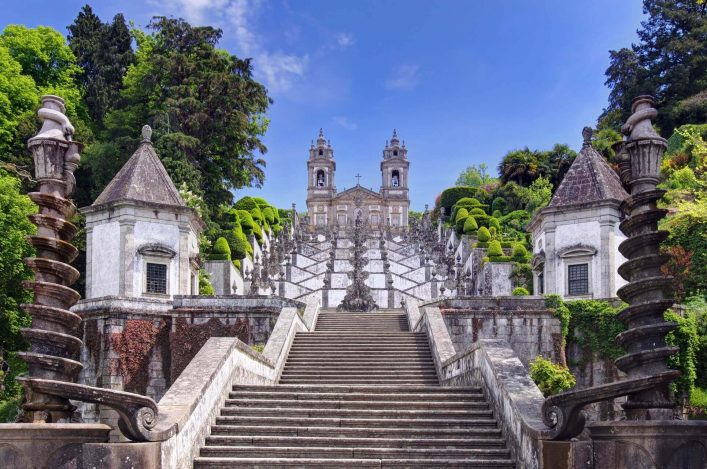 Breite Steintreppe führt zu einer barocken Kirche mit zwei Türmen. Symmetrische, dekorative Balustraden flankieren die Treppe. Üppiges Grün umrahmt das Bauwerk, klare, blaue Himmel im Hintergrund.