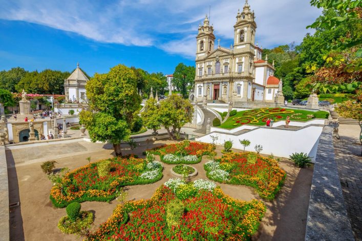 Bunte Gartenanlage mit Blumenbeeten in leuchtenden Rot- und Orangetönen vor der barocken Wallfahrtskirche, die auf einem Hügel steht. Eine geschwungene Treppe führt zur Kirche hinauf, Bäume umrahmen die Szenerie.