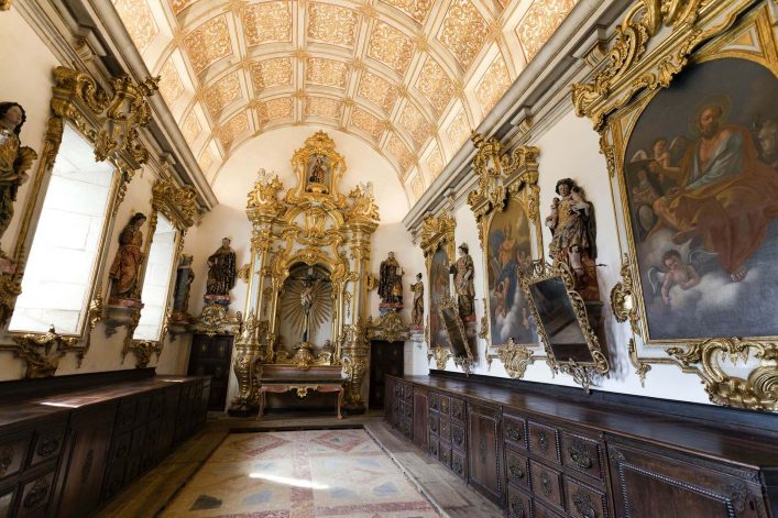 Prunkvoller Kirchensaal mit vergoldetem Altar in der Mitte, links und rechts umrahmt von Heiligenstatuen und Gemälden, Holzbänke entlang der Wände, helles Tageslicht fällt von links ein.