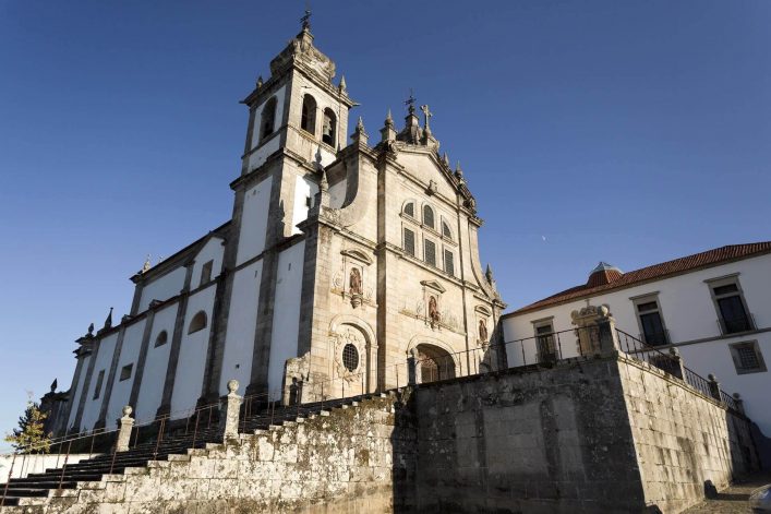 Prächtige Kirche mit steinernem, verziertem Glockenturm im Vordergrund, umgeben von einer Treppe und Mauer. Rechts ein kleineres, weißes Gebäude mit roten Ziegeln unter klarem blauen Himmel.