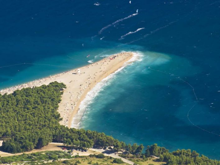 Ein goldener Sandstrand erstreckt sich als schmale Landzunge ins türkisblaue Meer. Umgeben von dichtem, grünem Wald im Vordergrund, während sich Menschen entlang des Strandes aufhalten.