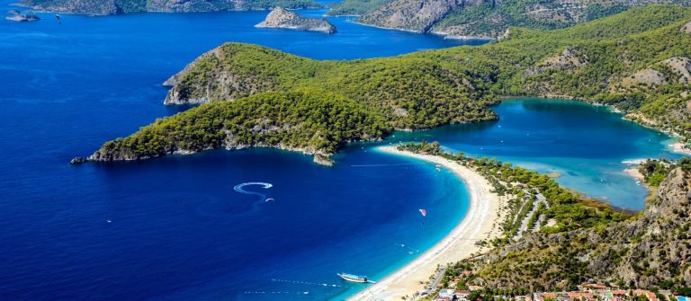 Strand mit feinem Sand erstreckt sich entlang der türkisblauen Küste. Im Hintergrund erheben sich grüne Hügel. Ein Paragleiter schwebt über dem Wasser. Boot am Ufer. Entspanntes Ferienparadies.