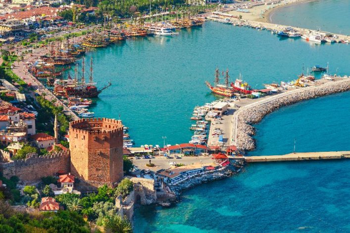 Roter Kizil Kule Turm in Alanya an der türkischen Riviera, umgeben von der historischen Festungsmauer, daneben blau schimmernder Hafen mit Booten, grün bewachsene Landschaft im Hintergrund.
